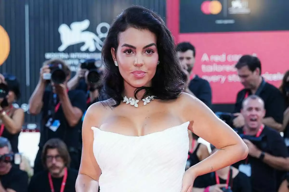 Mujer con vestido blanco elegante y collar de flores posando en una alfombra roja mientras fotógrafos la retratan al fondo Mujer con vestido blanco elegante y collar de flores posando en una alfombra roja mientras fotógrafos la retratan al fondo