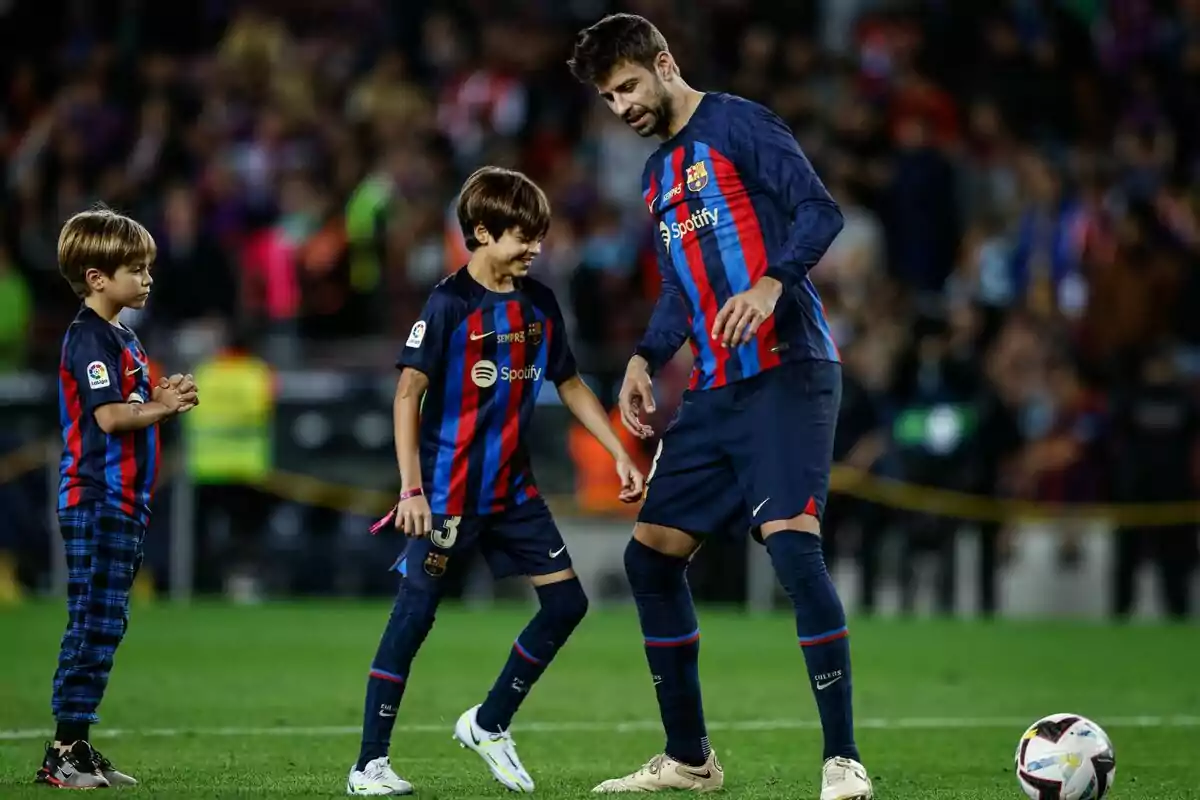 Un jugador de fútbol del FC Barcelona está en el campo con dos niños, todos vistiendo el uniforme del equipo, mientras interactúan y sonríen. Un jugador de fútbol del FC Barcelona está en el campo con dos niños, todos vistiendo el uniforme del equipo, mientras interactúan y sonríen.