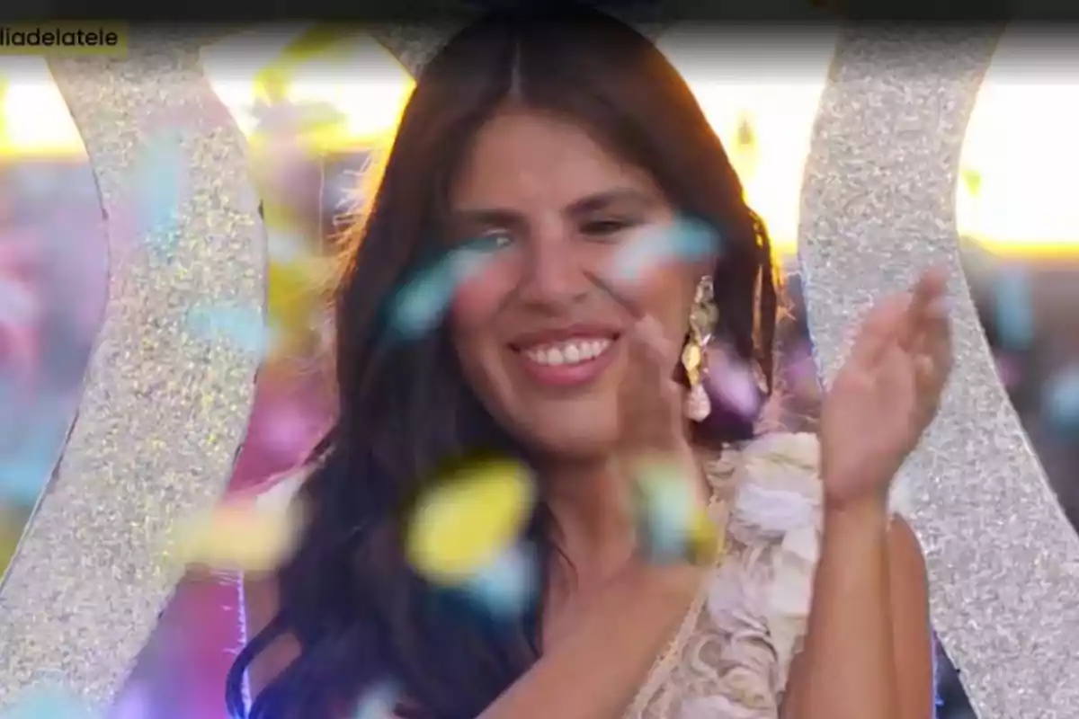 Isa Pantoja sonriente aplaude mientras confeti de colores cae a su alrededor en un ambiente festivo. Isa Pantoja sonriente aplaude mientras confeti de colores cae a su alrededor en un ambiente festivo.