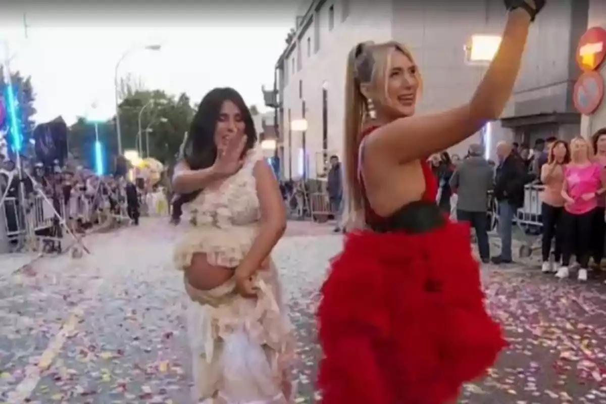 Isa Pantoja y Marta Riesco vestidas con trajes llamativos posan y sonríen en una calle llena de confeti mientras un grupo de personas observa detrás de una valla. Isa Pantoja y Marta Riesco vestidas con trajes llamativos posan y sonríen en una calle llena de confeti mientras un grupo de personas observa detrás de una valla.