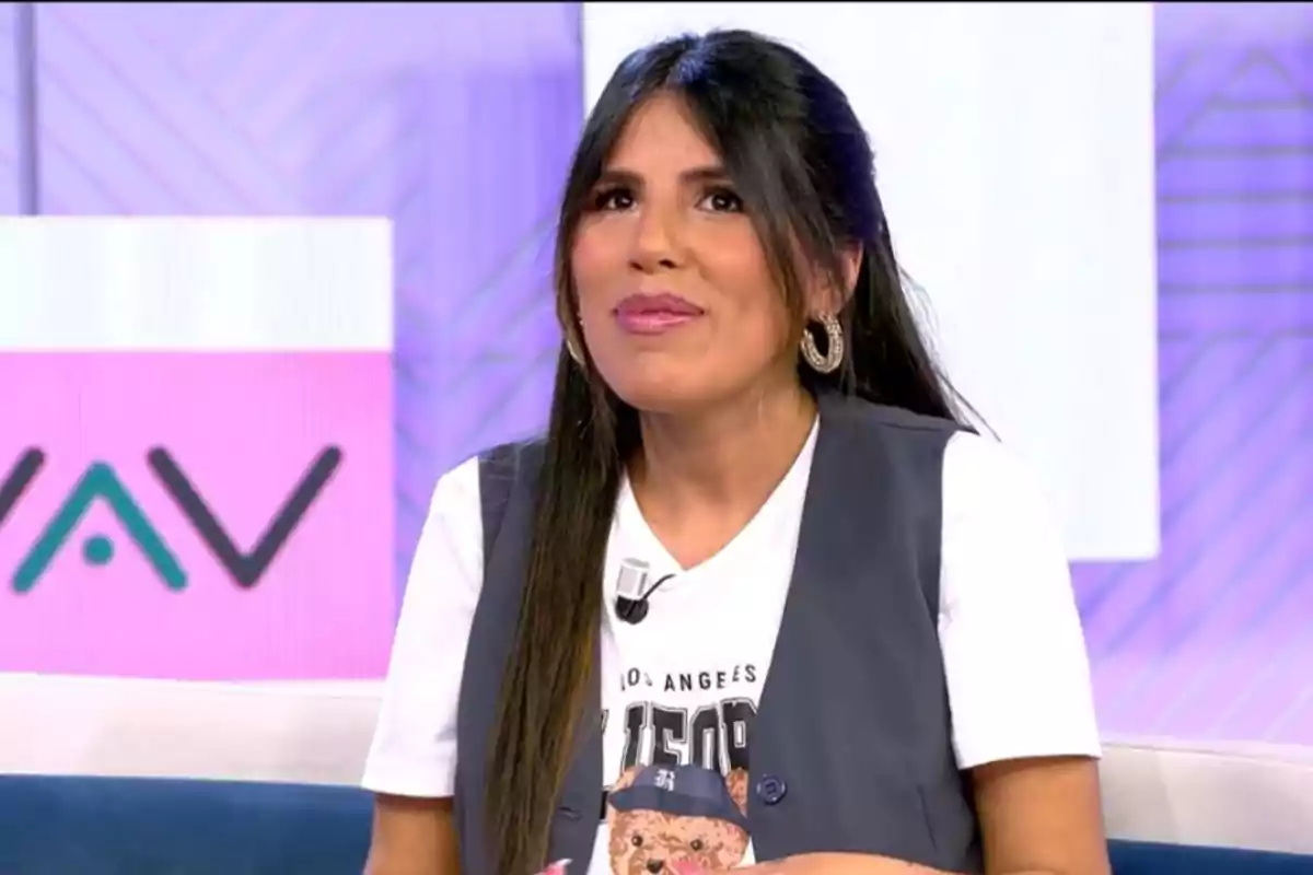 A woman with long dark hair is sitting on a television set with a colorful background, wearing a white T-jersey with a bear design and a gray vest, and she is smiling while looking up.