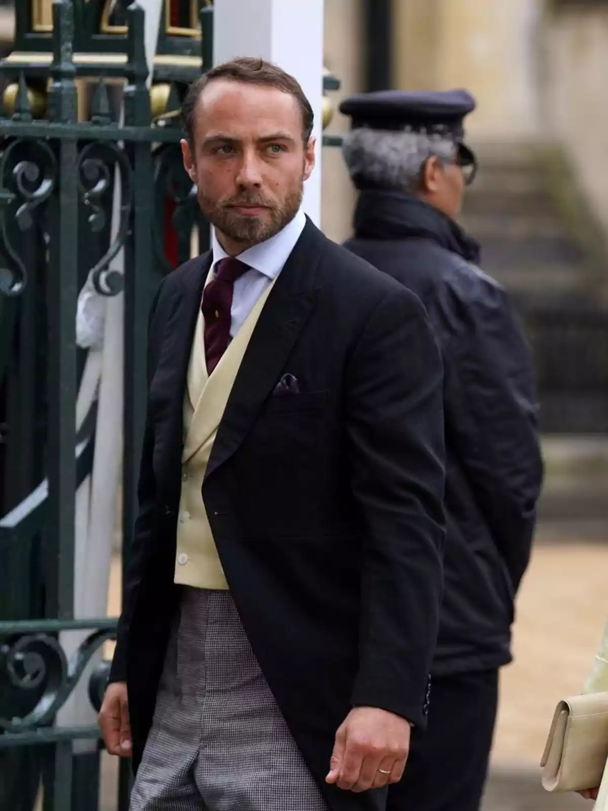 A bearded man with short hair, dressed in a formal suit with a black jacket, beige vest, red tie and checked trousers, is standing in front of a green fence, while a person in a dark uniform and cap stands with his back turned in the background. A bearded man with short hair, dressed in a formal suit with a black jacket, beige vest, red tie and checked trousers, is standing in front of a green fence, while a person in a dark uniform and cap stands with his back turned in the background.
