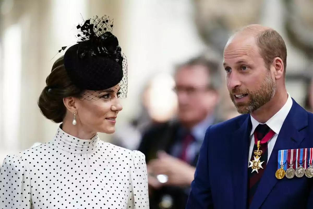 Una mujer con vestido blanco de lunares y sombrero negro conversa con un hombre de traje azul con medallas y condecoraciones en un evento formal Una mujer con vestido blanco de lunares y sombrero negro conversa con un hombre de traje azul con medallas y condecoraciones en un evento formal