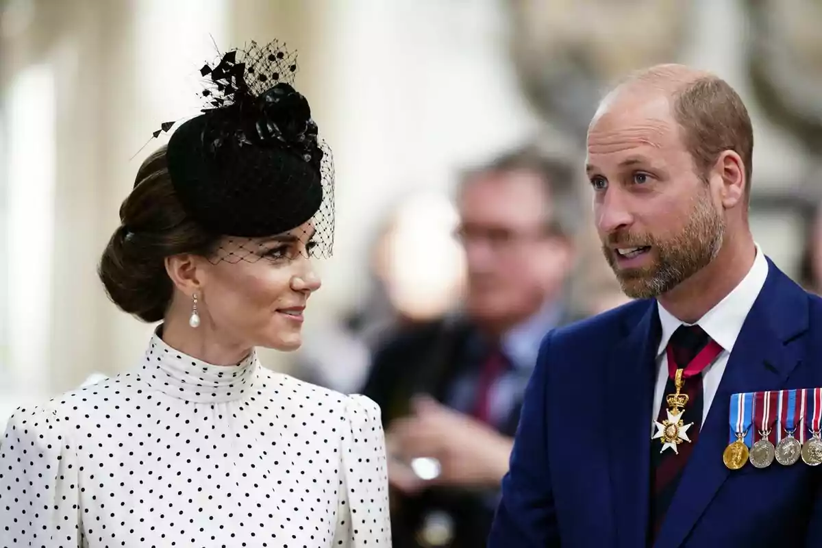 Una mujer con un elegante sombrero negro y vestido de lunares conversa con un hombre que lleva un traje azul y varias medallas. Una mujer con un elegante sombrero negro y vestido de lunares conversa con un hombre que lleva un traje azul y varias medallas.