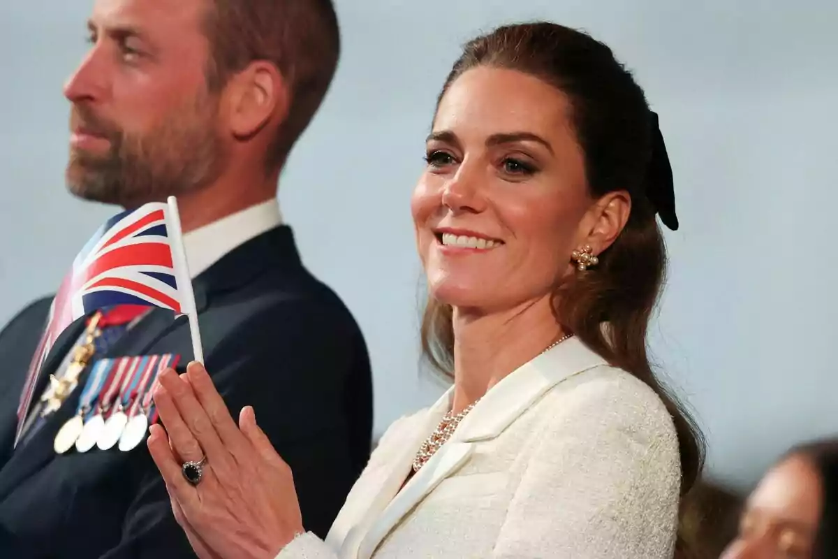 Una mujer sonriente sostiene una pequeña bandera del Reino Unido mientras está de pie junto a un hombre con medallas en su traje. Una mujer sonriente sostiene una pequeña bandera del Reino Unido mientras está de pie junto a un hombre con medallas en su traje.