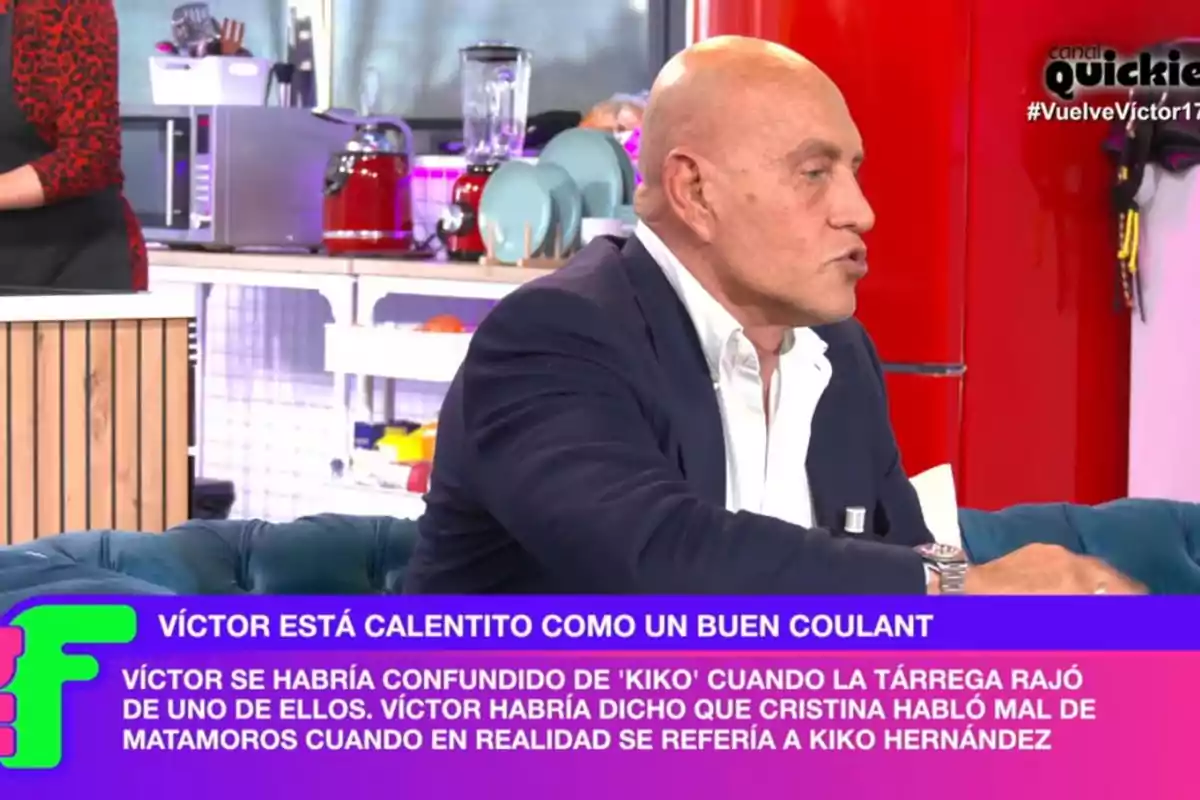 A bald man in a dark suit sits on a television set, while text appears on the screen at the bottom. A bald man in a dark suit sits on a television set, while text appears on the screen at the bottom.