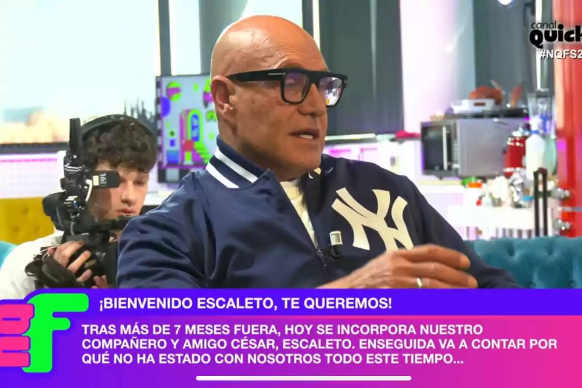 Kiko Matamoros with glasses and a blue jacket with the Yankees logo is speaking on 'Ni que fuéramos', while a cameraman records him; on the screen, there is a welcome message for a colleague named César, also known as Escaleto.