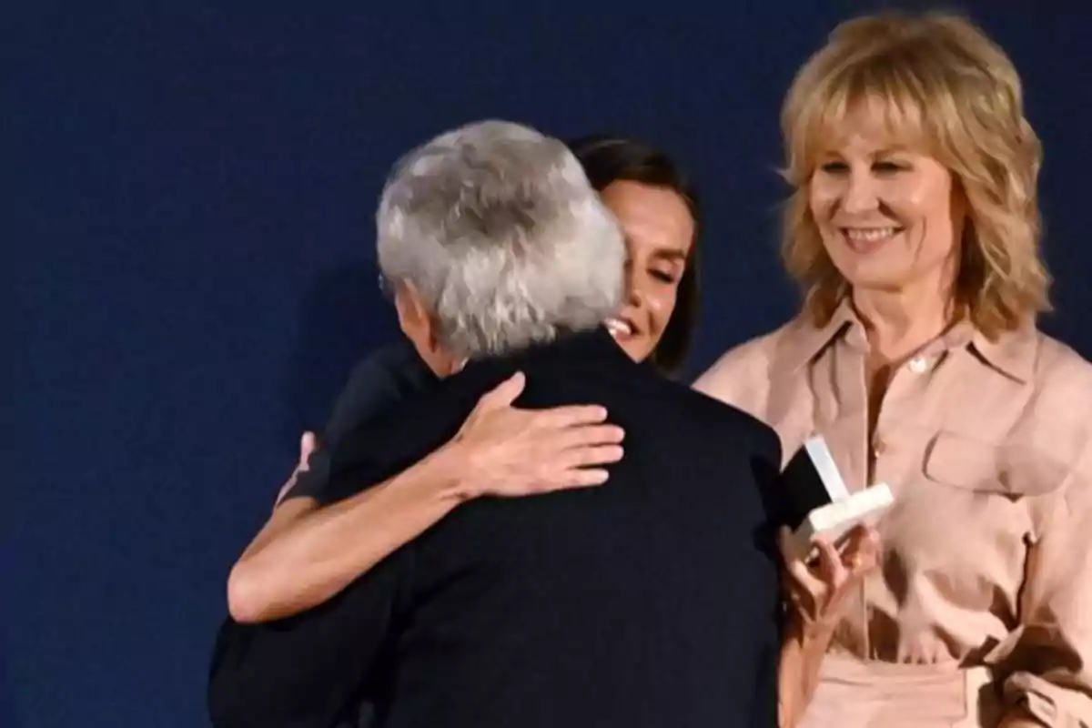 La reina Letizia abrazando a Fernando Ónega mayor mientras otra mujer sonríe sosteniendo una caja pequeña. La reina Letizia abrazando a Fernando Ónega mayor mientras otra mujer sonríe sosteniendo una caja pequeña.
