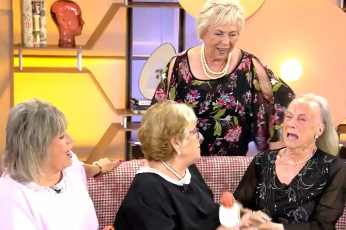 Un grupo de mujeres mayores está sentado en un sofá en un ambiente de estudio de televisión, una de ellas parece emocionada mientras las otras la observan y conversan. Un grupo de mujeres mayores está sentado en un sofá en un ambiente de estudio de televisión, una de ellas parece emocionada mientras las otras la observan y conversan.