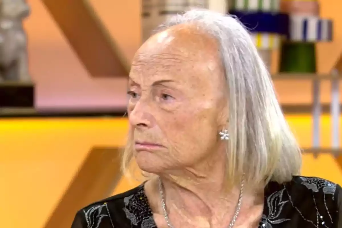Lola mayor con cabello canoso y expresión seria, usando aretes brillantes y un collar, en un entorno interior con fondo desenfocado. Lola mayor con cabello canoso y expresión seria, usando aretes brillantes y un collar, en un entorno interior con fondo desenfocado.