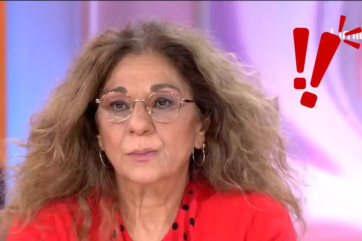 Lolita Flores con cabello rizado y gafas, vestida de rojo, aparece en un entorno de estudio con un gráfico de exclamación roja en la esquina.