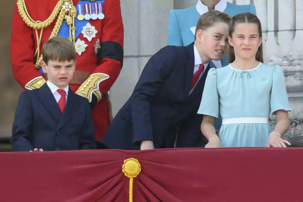Three children dressed formally watch an event from a balcony decorated with red fabric and gold details, while elegantly dressed adults stand behind them.