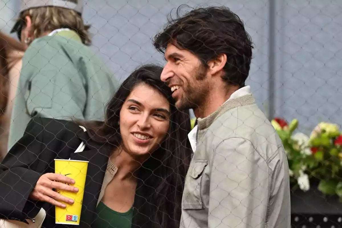 Lucía Rivera y Cayetano Rivera sonríen mientras observan un evento deportivo detrás de una malla. Lucía Rivera y Cayetano Rivera sonríen mientras observan un evento deportivo detrás de una malla.