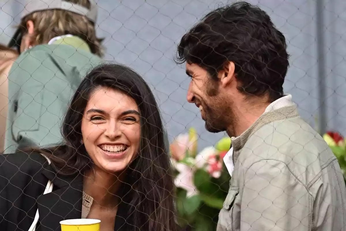 Lucía Rivera sonriente sostiene un vaso amarillo mientras conversa con Cayetano Rivera, ambos están detrás de una malla y parecen estar disfrutando de un evento al aire libre. Lucía Rivera sonriente sostiene un vaso amarillo mientras conversa con Cayetano Rivera, ambos están detrás de una malla y parecen estar disfrutando de un evento al aire libre.