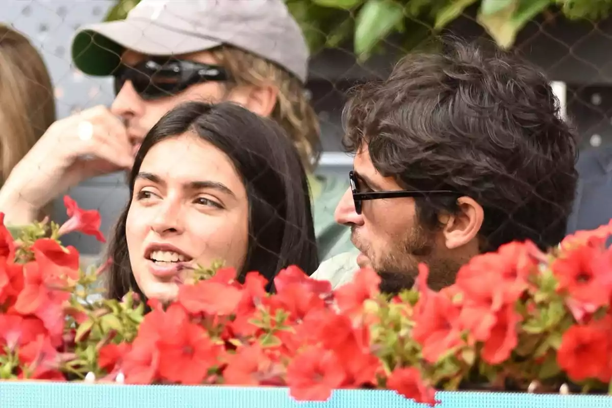 Un grupo de personas está sentado detrás de una fila de flores rojas, conversando y disfrutando de un evento al aire libre. Un grupo de personas está sentado detrás de una fila de flores rojas, conversando y disfrutando de un evento al aire libre.