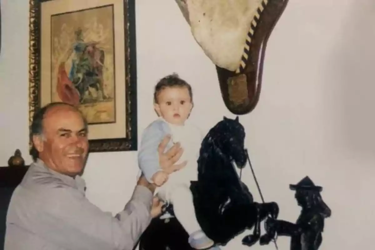 Hombre sonriente sostiene a un bebé sentado sobre una escultura de caballo negro en una habitación decorada con un cuadro y un objeto en la pared Hombre sonriente sostiene a un bebé sentado sobre una escultura de caballo negro en una habitación decorada con un cuadro y un objeto en la pared