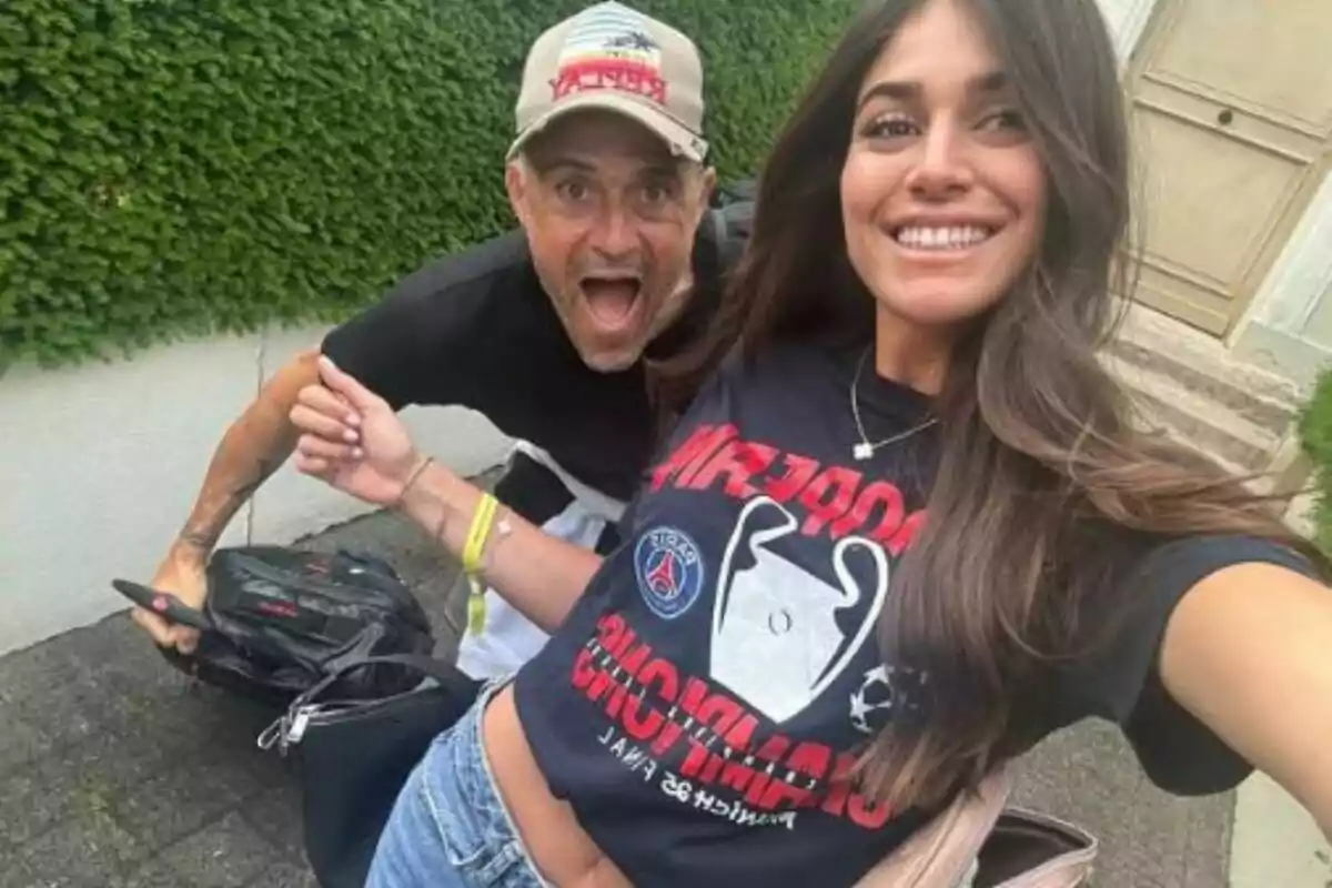 Una joven sonriente toma una selfie junto a un hombre que hace un gesto divertido al aire libre frente a una pared cubierta de plantas Una joven sonriente toma una selfie junto a un hombre que hace un gesto divertido al aire libre frente a una pared cubierta de plantas
