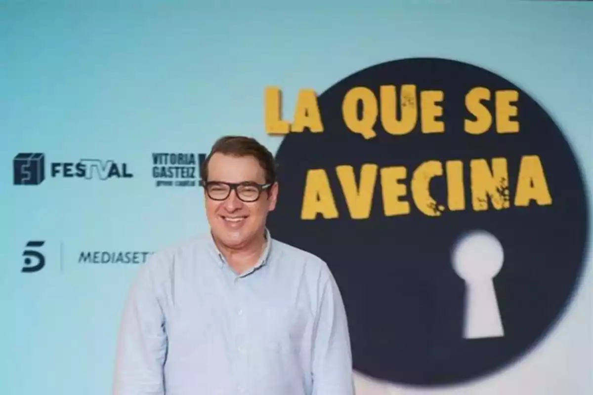 Luis Merlo con gafas sonríe frente a un cartel de "La que se avecina" en un evento de Mediaset.