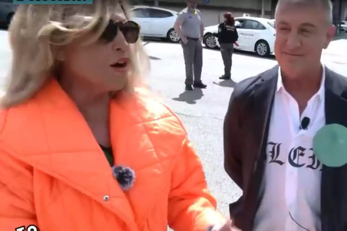 Lydia Lozano y Victor Sandoval conversan al aire libre Lydia lleva una chaqueta naranja brillante y gafas de sol mientras que Víctor viste una chaqueta oscura y una camisa blanca con letras. Lydia Lozano y Victor Sandoval conversan al aire libre Lydia lleva una chaqueta naranja brillante y gafas de sol mientras que Víctor viste una chaqueta oscura y una camisa blanca con letras.