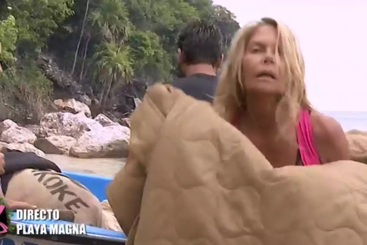 Makoke rubia con camiseta rosa sostiene una manta beige en la orilla de una playa con vegetación y rocas al fondo mientras otra persona está de espaldas y se ven sacos en una lancha azul Makoke rubia con camiseta rosa sostiene una manta beige en la orilla de una playa con vegetación y rocas al fondo mientras otra persona está de espaldas y se ven sacos en una lancha azul