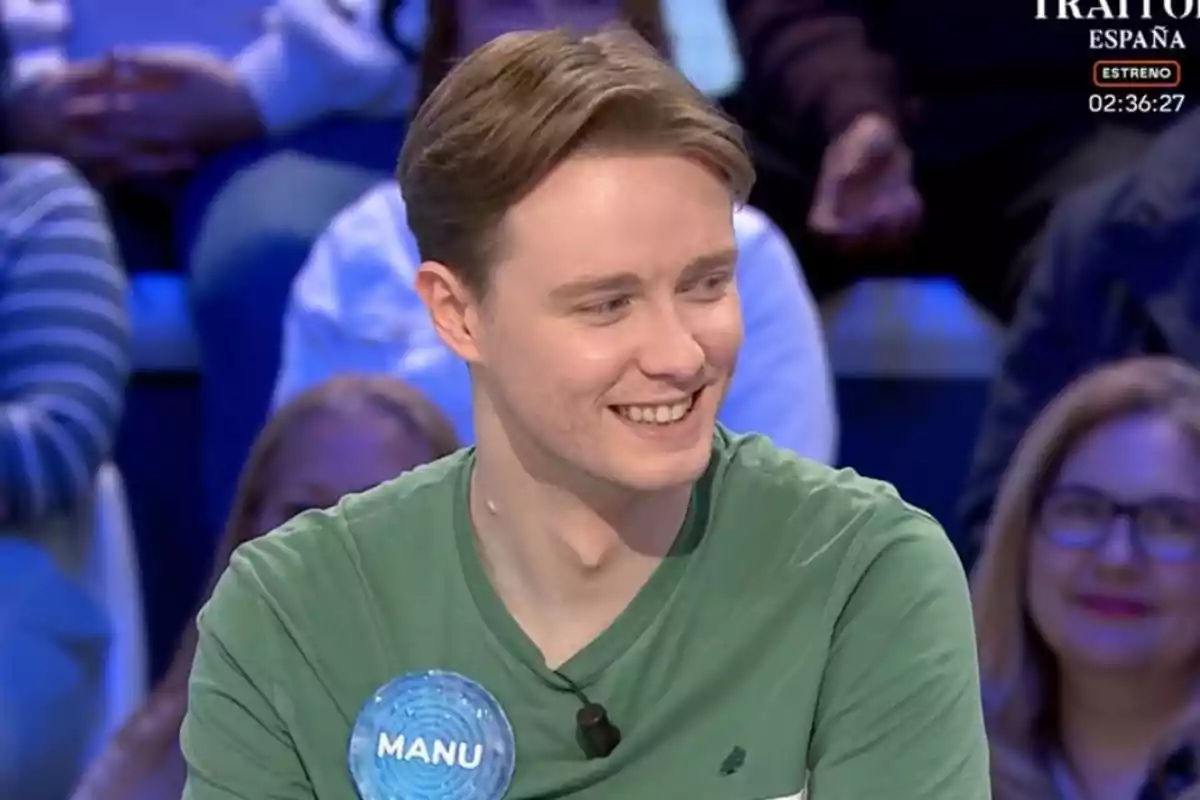 Manu joven con camiseta verde sonríe mientras está sentado en un programa de televisión, con un público desenfocado al fondo. Manu joven con camiseta verde sonríe mientras está sentado en un programa de televisión, con un público desenfocado al fondo.