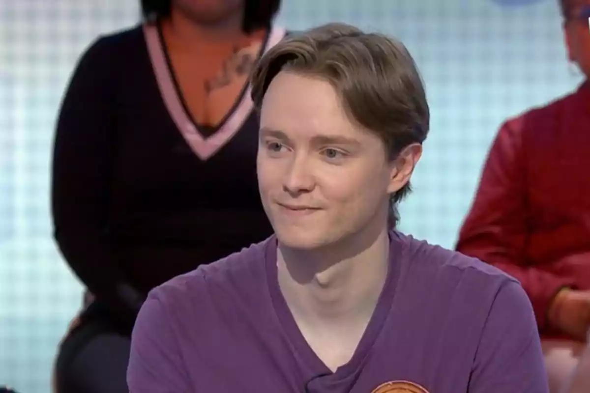 Manu joven con cabello castaño claro y camiseta morada está sentado en un entorno de estudio de televisión, con personas desenfocadas en el fondo. Manu joven con cabello castaño claro y camiseta morada está sentado en un entorno de estudio de televisión, con personas desenfocadas en el fondo.