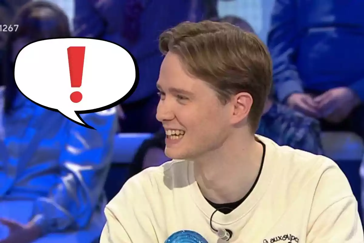 Manu joven sonríe mientras está sentado en un programa de televisión, con un globo de diálogo que contiene un signo de exclamación.