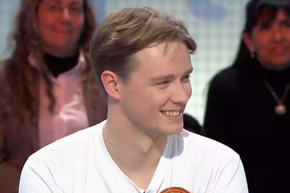 Manu joven sonríe mientras está sentado en un entorno que parece ser un programa de televisión, con personas en el fondo observando. Manu joven sonríe mientras está sentado en un entorno que parece ser un programa de televisión, con personas en el fondo observando.