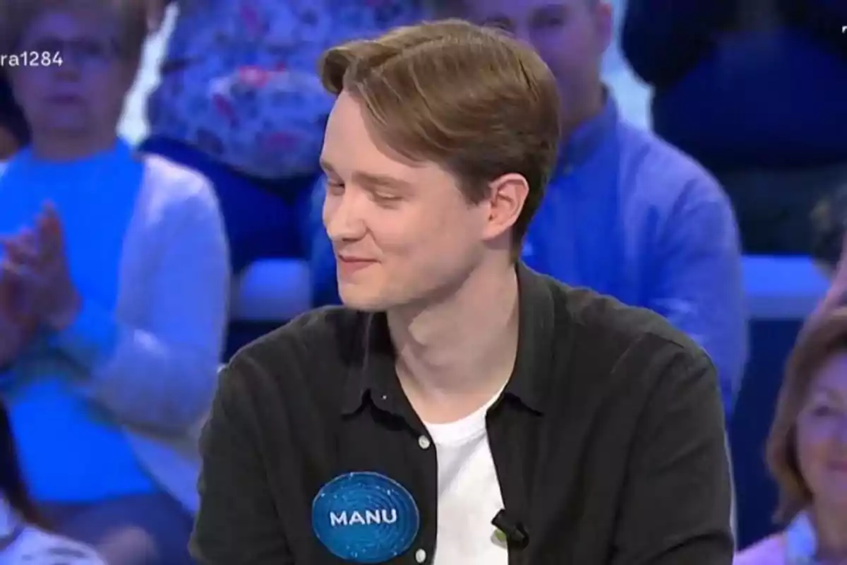 Manu con cabello castaño claro y camisa negra sonríe mientras participa en un programa de televisión, lleva una etiqueta azul con el nombre Manu y hay público desenfocado al fondo. Manu con cabello castaño claro y camisa negra sonríe mientras participa en un programa de televisión, lleva una etiqueta azul con el nombre Manu y hay público desenfocado al fondo.