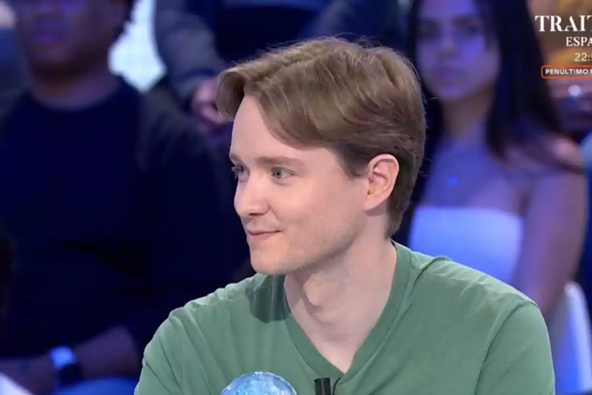 Manu de cabello castaño claro y camiseta verde sentado en un estudio de televisión con personas desenfocadas al fondo Manu de cabello castaño claro y camiseta verde sentado en un estudio de televisión con personas desenfocadas al fondo