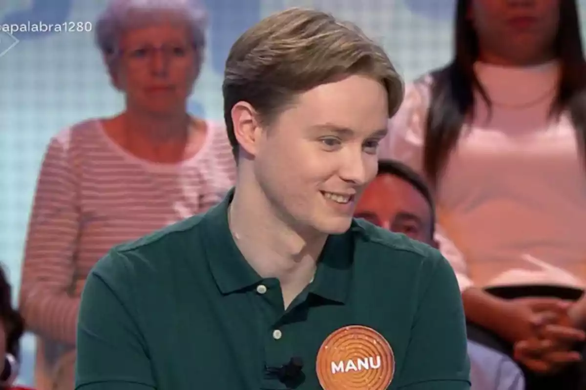 Manu con camisa verde y una placa que dice Manu participando en un programa de televisión con público de fondo Manu con camisa verde y una placa que dice Manu participando en un programa de televisión con público de fondo