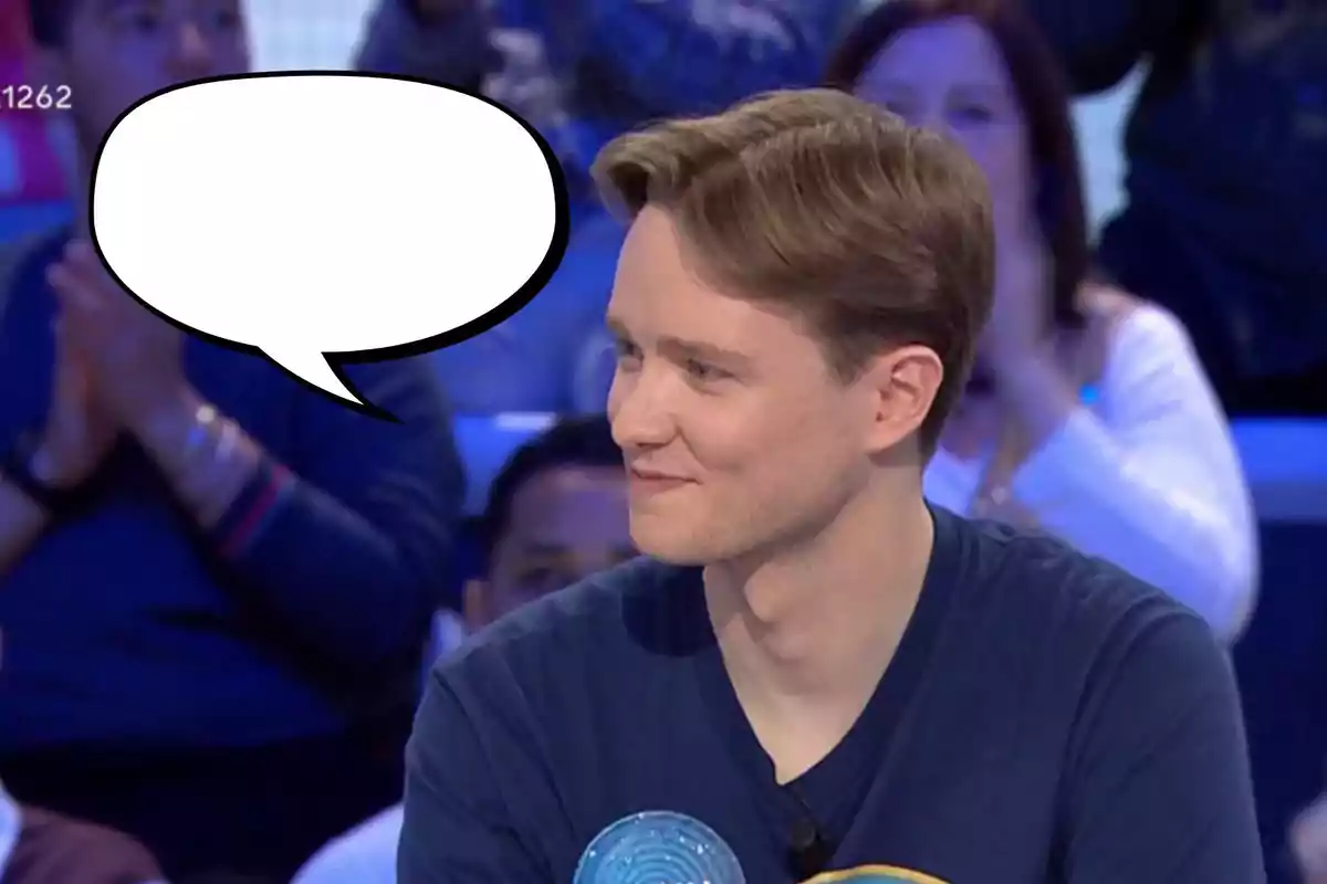 Manu joven con cabello claro y camiseta oscura está sentado en un estudio de televisión, sonriendo mientras mira hacia un lado; hay un globo de diálogo vacío sobre su cabeza y personas desenfocadas en el fondo.