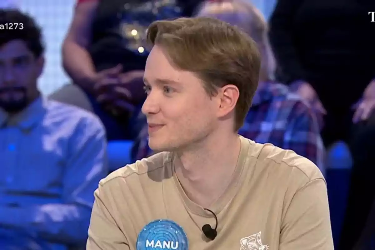 Manu joven con cabello claro y camiseta beige está sentado en un estudio de televisión con un micrófono en la solapa y una placa que dice Manu joven con cabello claro y camiseta beige está sentado en un estudio de televisión con un micrófono en la solapa y una placa que dice