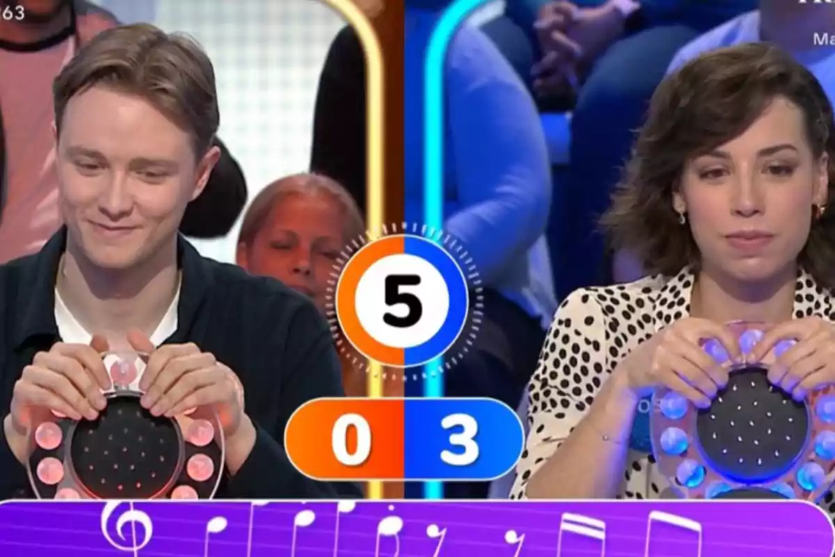 Rosa y Manu participan en un concurso de preguntas y respuestas, cada una sosteniendo un dispositivo de juego, con un marcador que muestra un puntaje de 0 a 3 y un temporizador en el centro. Rosa y Manu participan en un concurso de preguntas y respuestas, cada una sosteniendo un dispositivo de juego, con un marcador que muestra un puntaje de 0 a 3 y un temporizador en el centro.