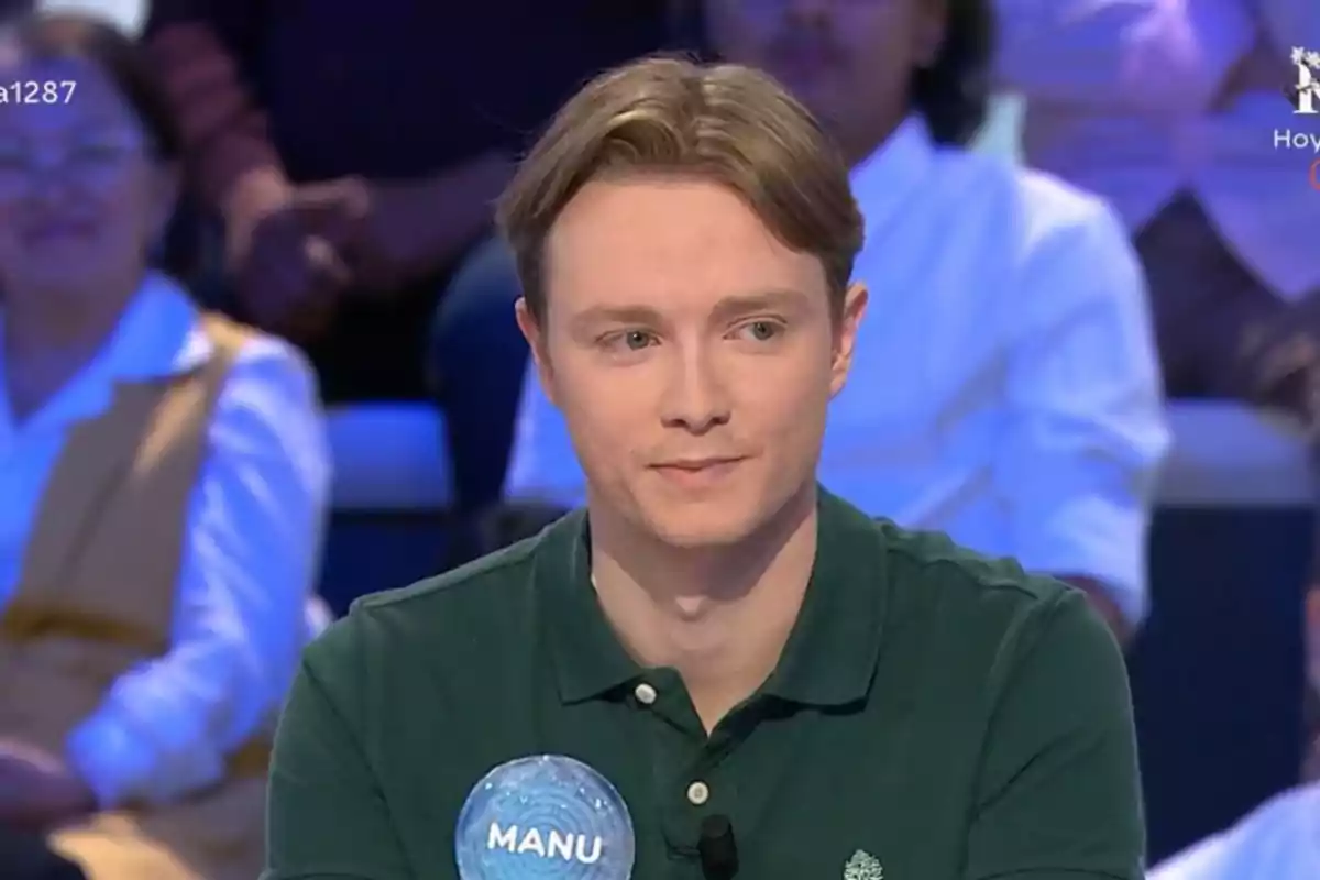 Manu de cabello rubio y camisa verde sentado en un programa de concursos con público al fondo y una etiqueta que dice Manu en su pecho Manu de cabello rubio y camisa verde sentado en un programa de concursos con público al fondo y una etiqueta que dice Manu en su pecho