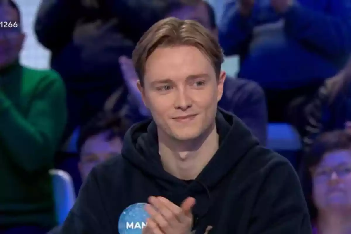 Manu con cabello claro y sudadera oscura aplaude mientras está sentado en un lugar con público al fondo. Manu con cabello claro y sudadera oscura aplaude mientras está sentado en un lugar con público al fondo.