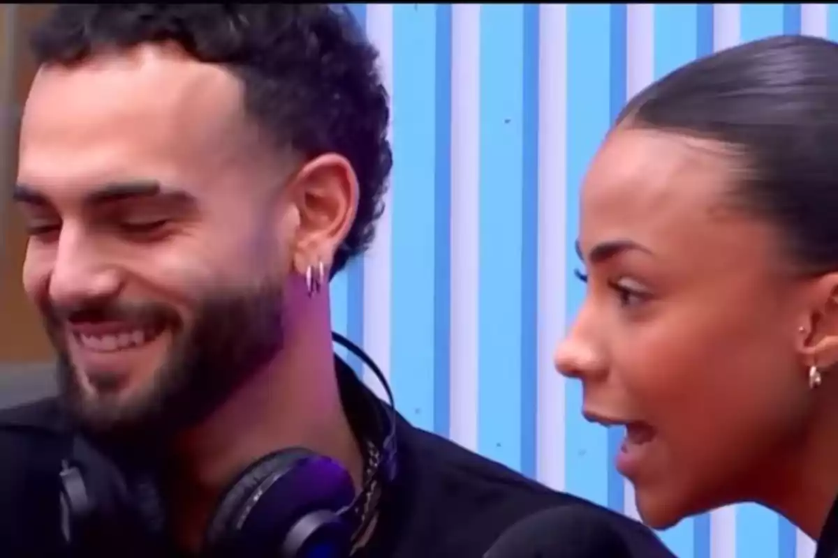 Manuel Cortés y Jeimy Báez conversando y sonriendo en un entorno con fondo de rayas azules. Manuel Cortés y Jeimy Báez conversando y sonriendo en un entorno con fondo de rayas azules.
