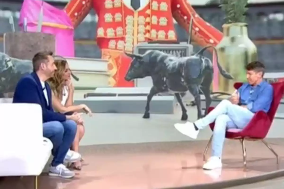 Manuel Díaz ‘El Cordobés’, Frank Blanco y Verónica Dulanto conversan en un set de televisión con una decoración que incluye una figura de un toro y un fondo con un traje de torero. Manuel Díaz ‘El Cordobés’, Frank Blanco y Verónica Dulanto conversan en un set de televisión con una decoración que incluye una figura de un toro y un fondo con un traje de torero.