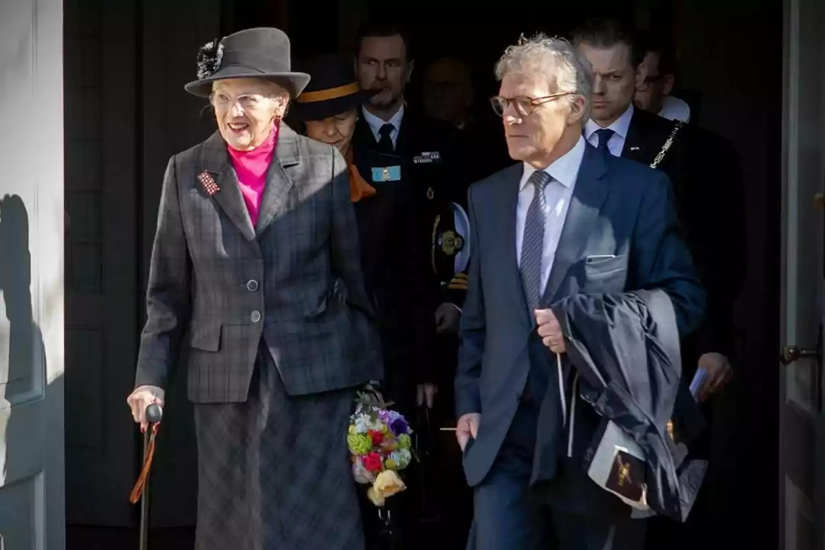 An elderly woman with a hat and cane walks alongside a man in a suit as they leave a building accompanied by other people. An elderly woman with a hat and cane walks alongside a man in a suit as they leave a building accompanied by other people.