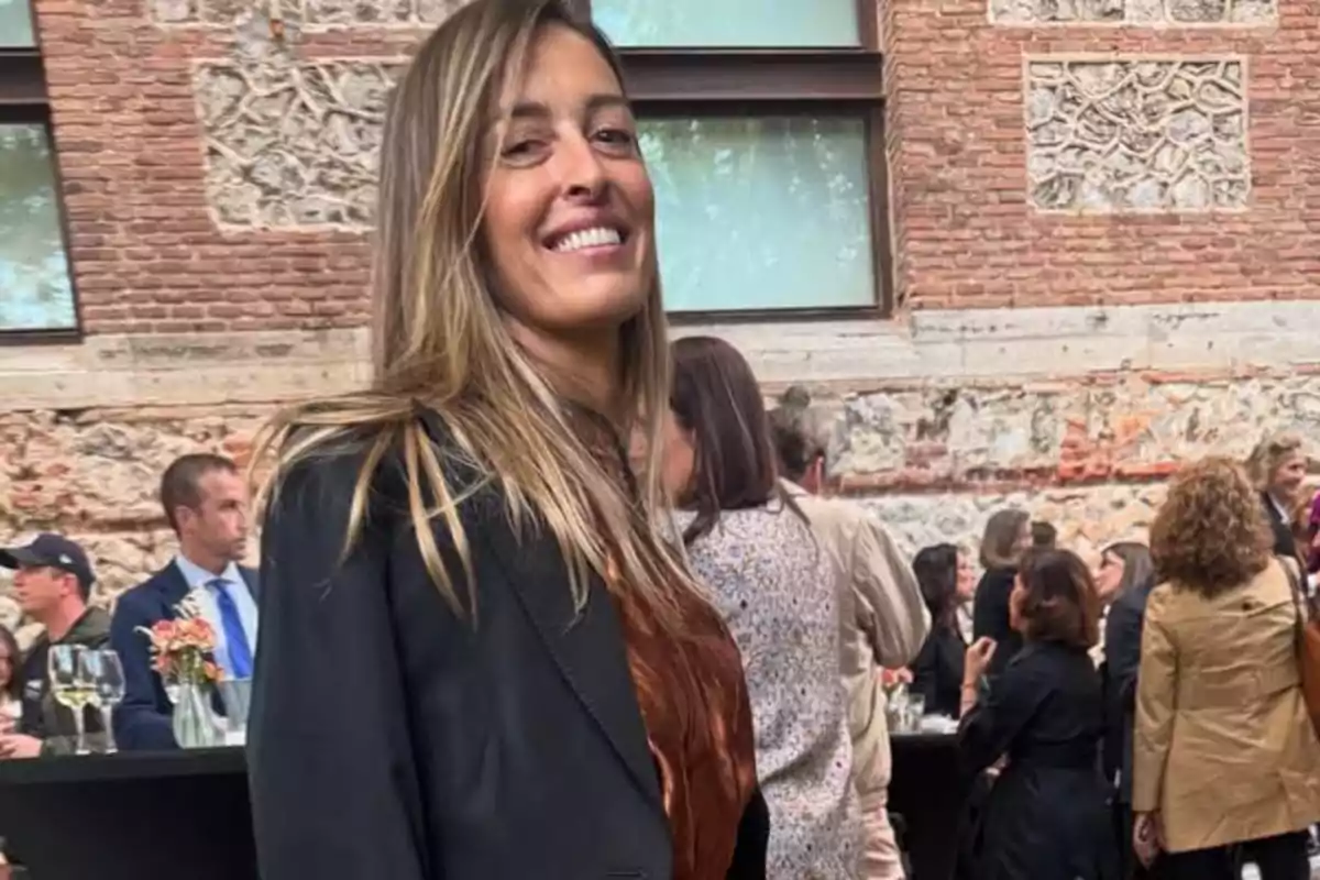 María Amores sonriente con cabello largo y rubio está de pie en un evento social al aire libre, rodeada de personas conversando y disfrutando de bebidas, con un edificio de ladrillo al fondo. María Amores sonriente con cabello largo y rubio está de pie en un evento social al aire libre, rodeada de personas conversando y disfrutando de bebidas, con un edificio de ladrillo al fondo.