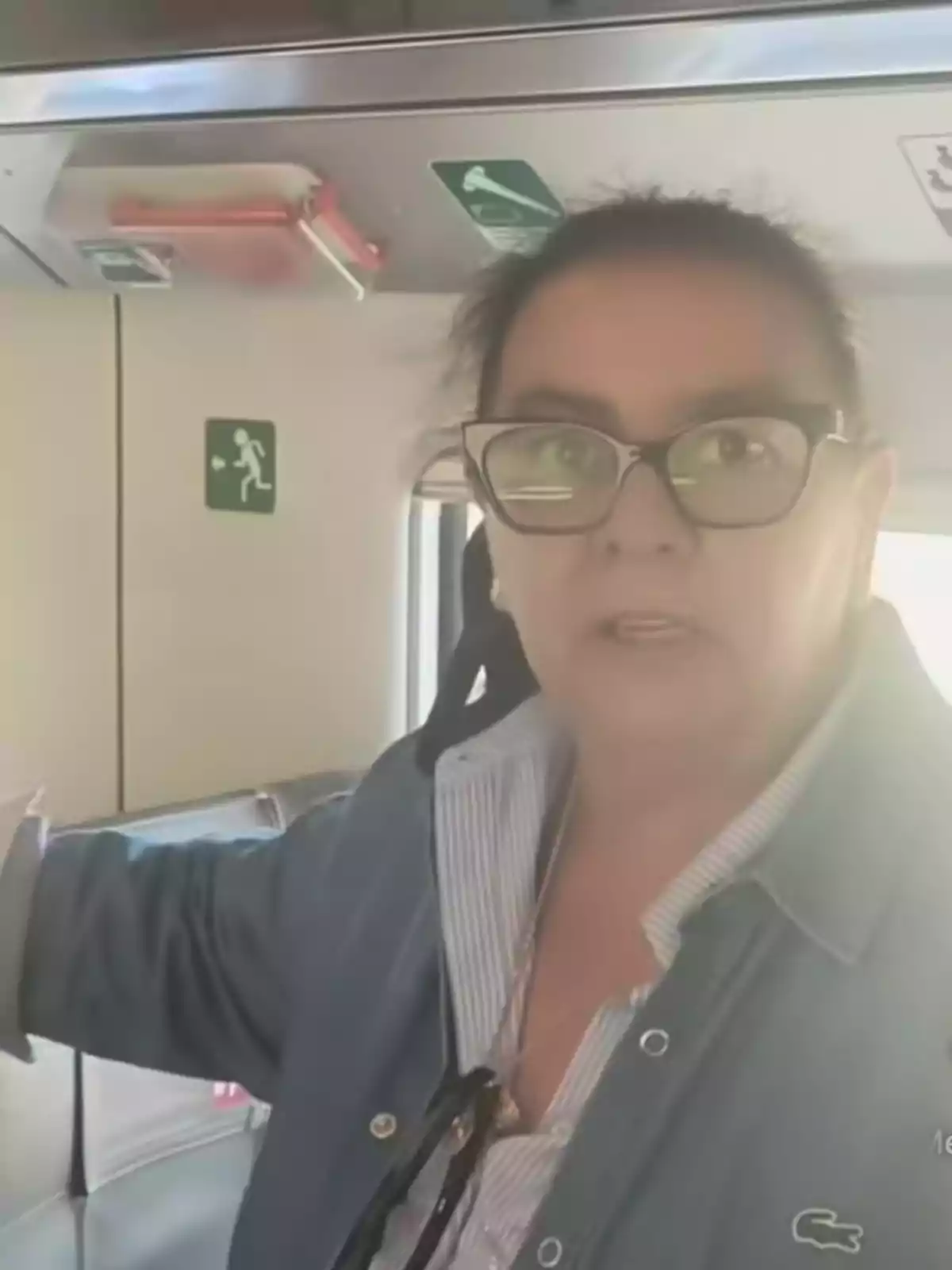 María del Monte con gafas y chaqueta azul está de pie en el interior de un tren, junto a señales de emergencia en la pared. María del Monte con gafas y chaqueta azul está de pie en el interior de un tren, junto a señales de emergencia en la pared.