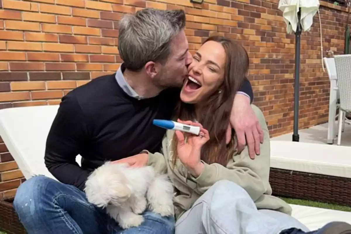 Marta Peñate y Tony Spina sonrientes se abraza mientras la mujer sostiene una prueba de embarazo y un pequeño perro blanco está en su regazo. Marta Peñate y Tony Spina sonrientes se abraza mientras la mujer sostiene una prueba de embarazo y un pequeño perro blanco está en su regazo.