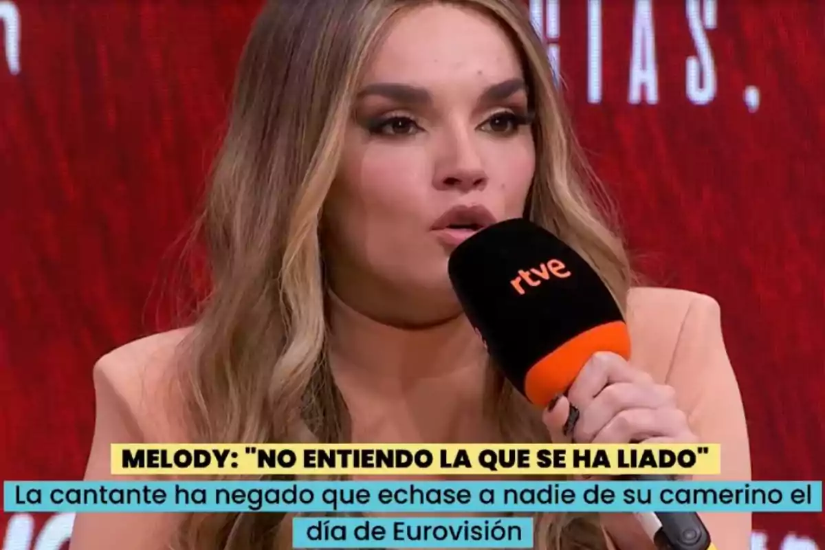 Melody con el cabello largo y rubio hablando en una entrevista mientras sostiene un micrófono negro con el logo de rtve, sobre un fondo rojo y con subtítulos en la parte inferior Melody con el cabello largo y rubio hablando en una entrevista mientras sostiene un micrófono negro con el logo de rtve, sobre un fondo rojo y con subtítulos en la parte inferior