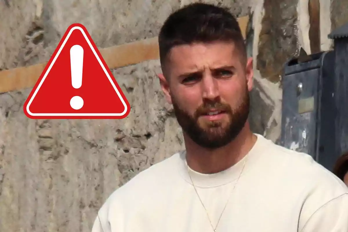 David Rodríguez con barba y cabello corto está de pie frente a una pared de piedra junto a un símbolo de advertencia rojo con un signo de exclamación blanco.