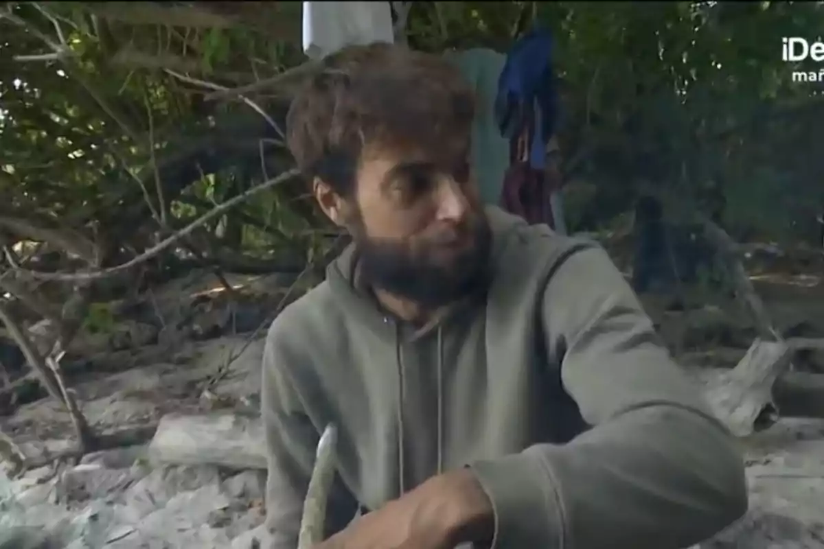 Montoya con barba y sudadera está sentado en un entorno natural rodeado de ramas y vegetación. Montoya con barba y sudadera está sentado en un entorno natural rodeado de ramas y vegetación.