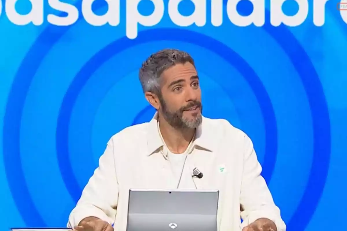 Roberto Leal de cabello corto y barba sentado frente a una computadora en un set de televisión con fondo azul y letras blancas Roberto Leal de cabello corto y barba sentado frente a una computadora en un set de televisión con fondo azul y letras blancas