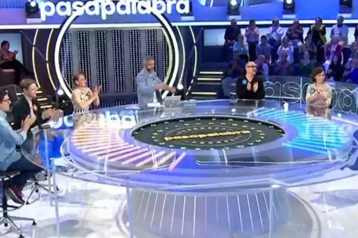 Un grupo de personas aplaude en un set de televisión con un diseño circular y un público al fondo. Un grupo de personas aplaude en un set de televisión con un diseño circular y un público al fondo.