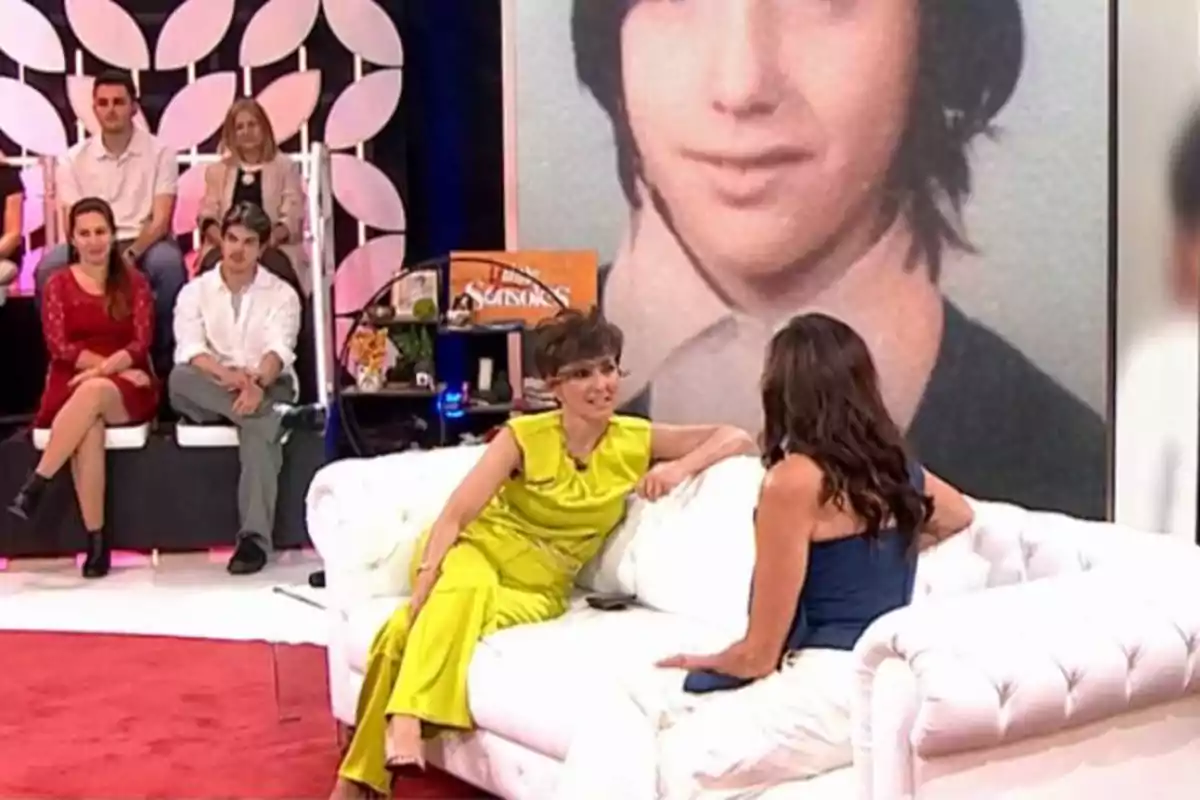 Un grupo de personas está en un set de televisión, dos mujeres conversan en un sofá blanco mientras otras personas están sentadas al fondo, y una gran imagen de una persona joven se muestra en la pantalla detrás de ellas. Un grupo de personas está en un set de televisión, dos mujeres conversan en un sofá blanco mientras otras personas están sentadas al fondo, y una gran imagen de una persona joven se muestra en la pantalla detrás de ellas.