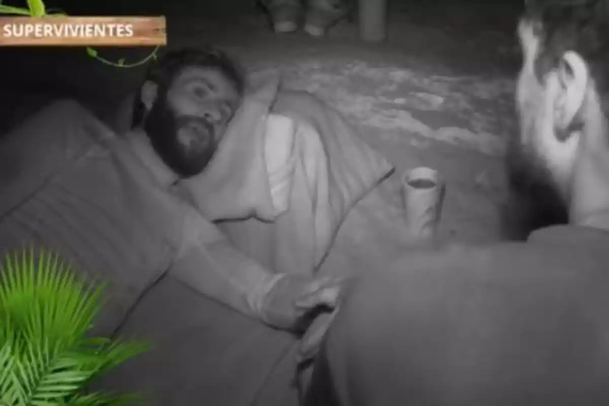 Pelayo y Joshua conversan en un entorno oscuro mientras Pelayo está recostado sobre una almohada, con un vaso al lado y un letrero que dice Pelayo y Joshua conversan en un entorno oscuro mientras Pelayo está recostado sobre una almohada, con un vaso al lado y un letrero que dice