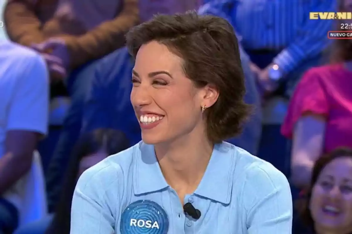 Rosa sonriente con cabello corto y camisa azul claro participando en un programa de televisión con público de fondo y una placa con el nombre Rosa en el pecho Rosa sonriente con cabello corto y camisa azul claro participando en un programa de televisión con público de fondo y una placa con el nombre Rosa en el pecho